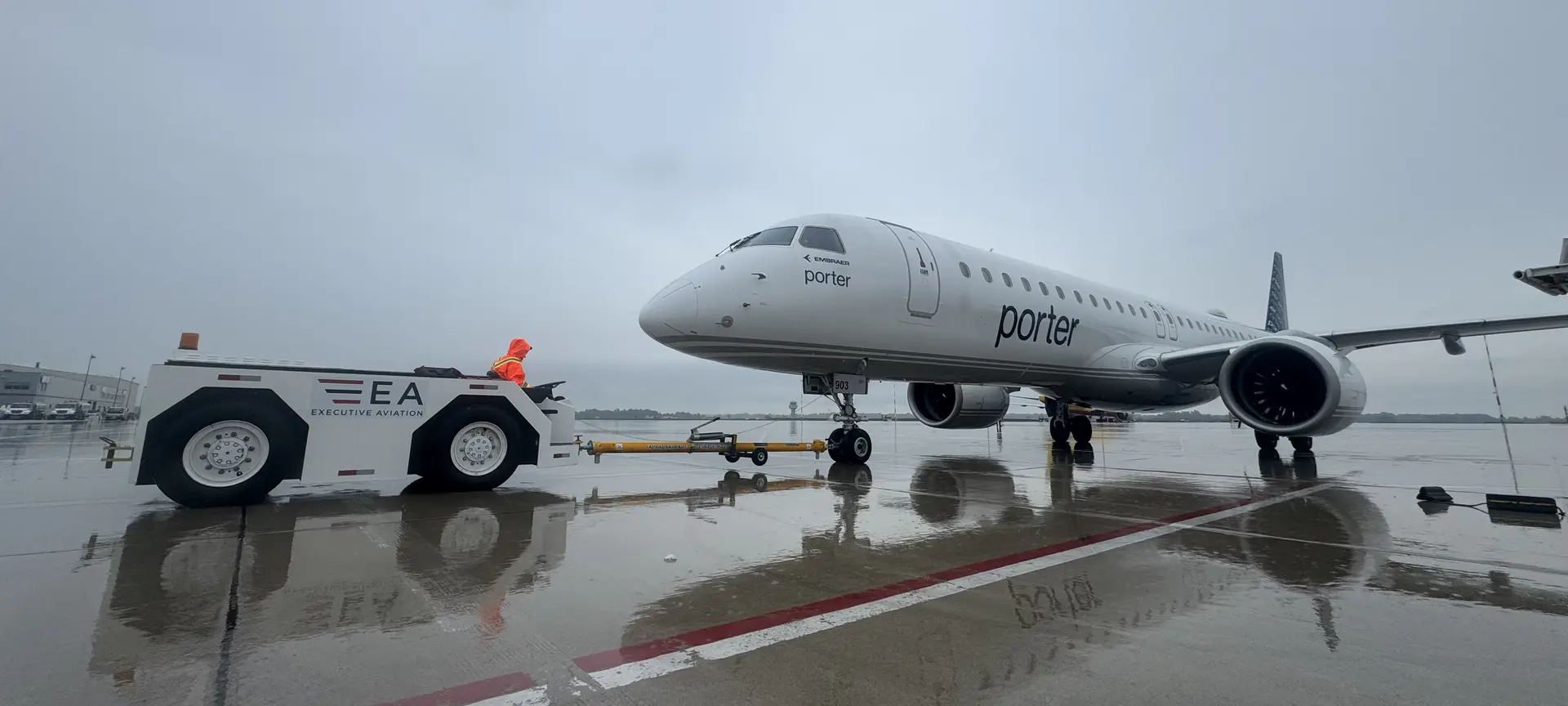 An enedym-converted electric pushback tractor towing a Porter Embraer E195 across a wet apron